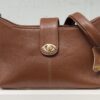 Brown pebbled leather handbag with a gold turn-lock closure and a long shoulder strap on a white surface.